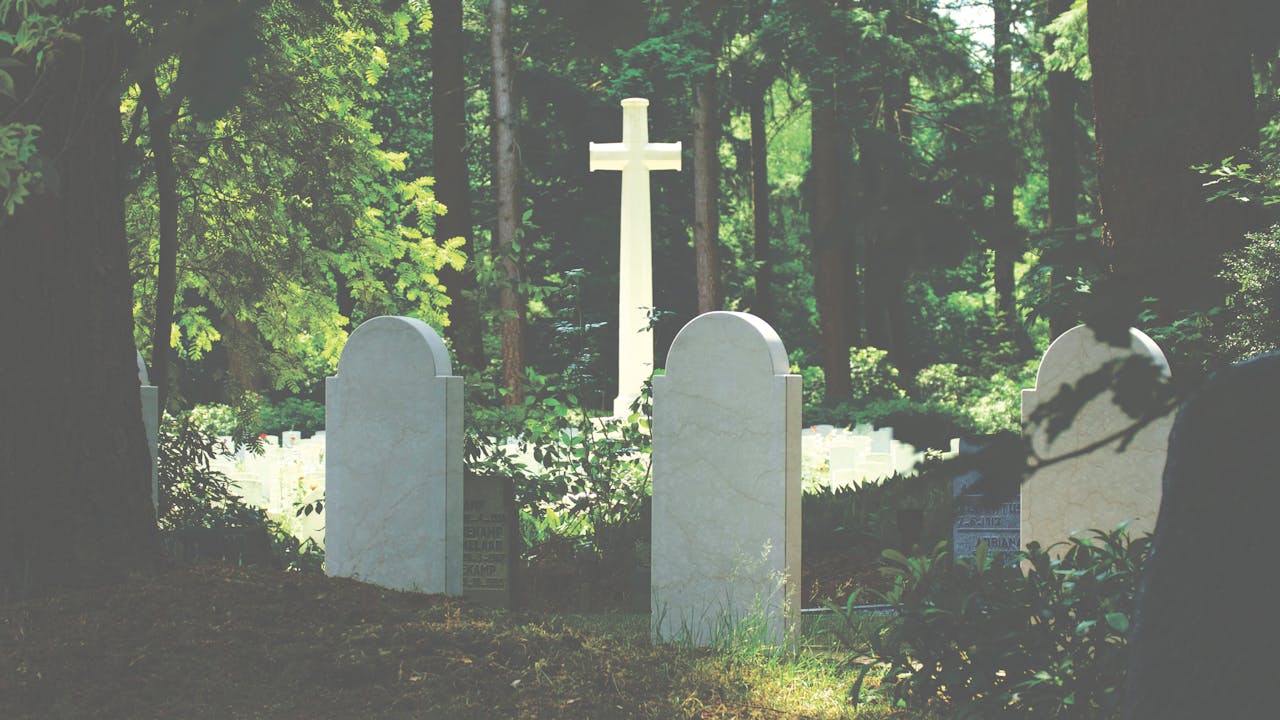 Peaceful cemetery scene with gravestones and cross amidst lush trees, evoking a sense of tranquility and reflection.